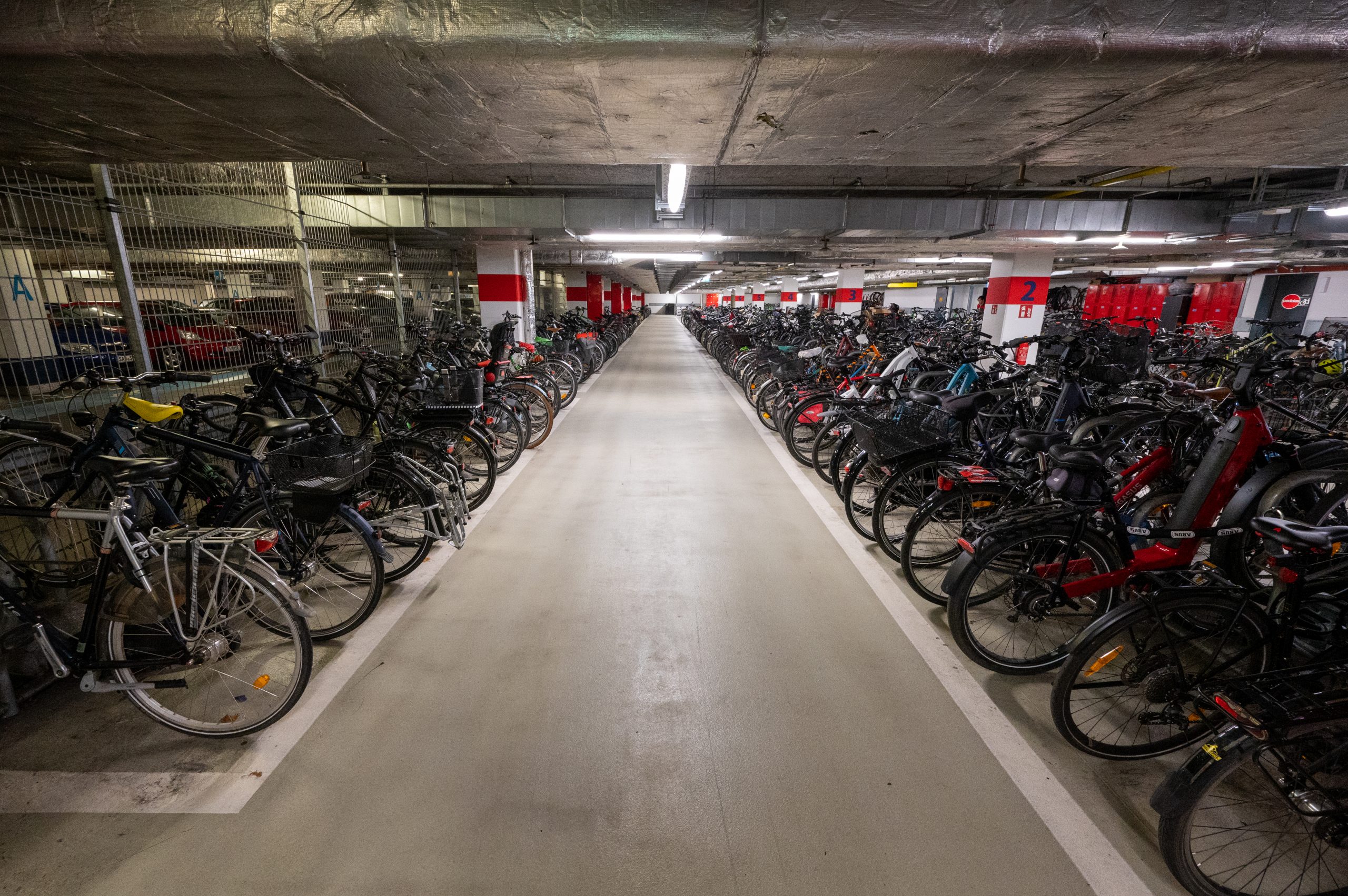 Blick in Reihen von geparkten Fahrrädern in Hoch-Tief-Parkern in einem Fahrradparkhaus, das durch einen Zaun von dem Bereich für PKW-Parken der Tiefgarage abgegrenzt ist.