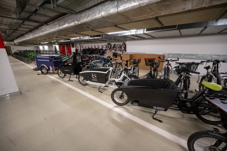 Blick auf schrägt geparkte Lastenräder in dafür gekennzeichneten Flächen in einer Tiefgarage. Im Hintergrund stehen sauber aufgereihte Leihfahrräder und an der Wand hängen zusammengerollte Fahrradschläuche