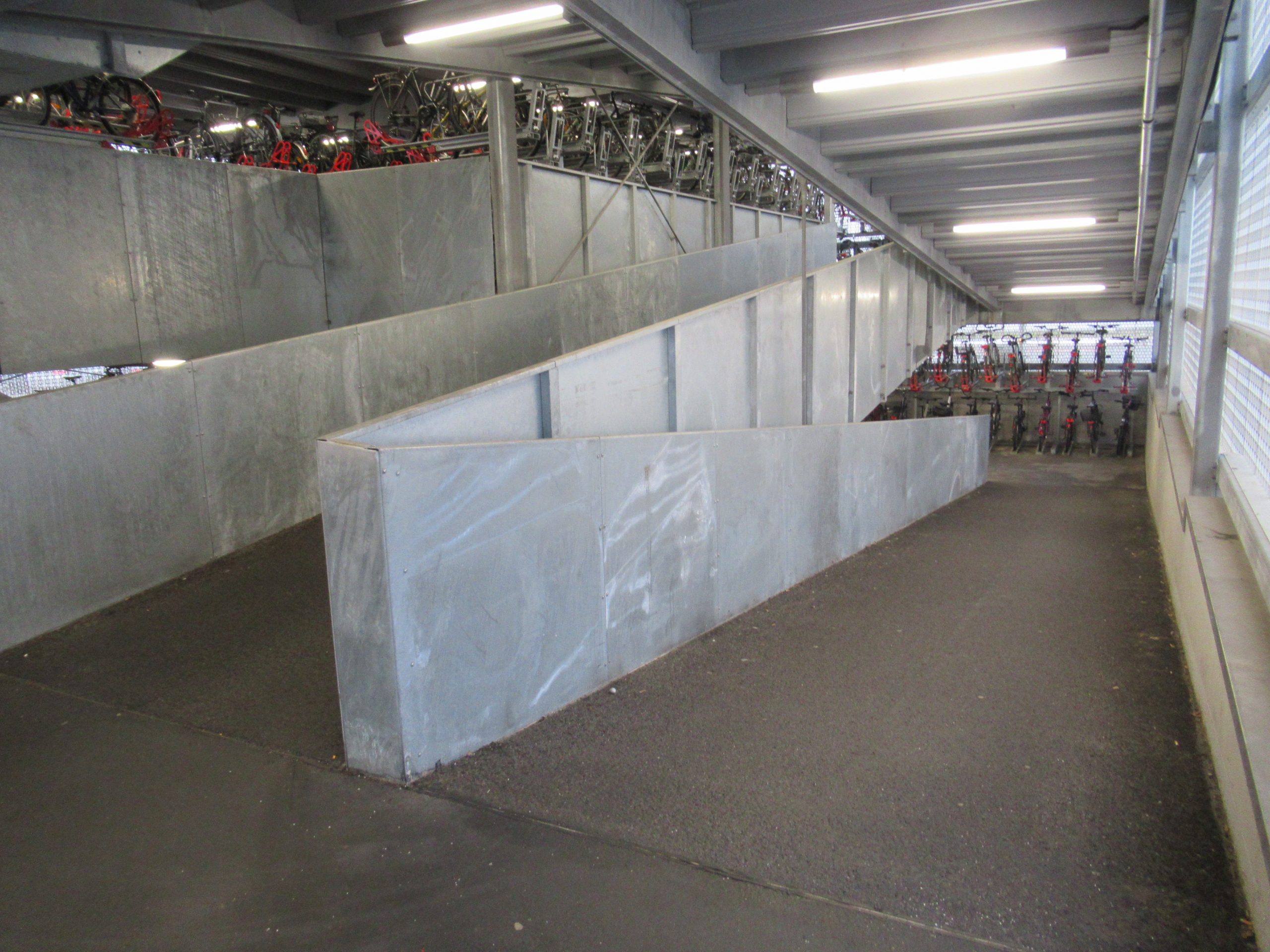 Fahrradrampe im Inneren des Fahrradparkhaus verbindet die Ebenen im Zick-Zack miteinander. Grauer Bodenbelag und Beton-Wände mit einer Höhe von ca. 110cm