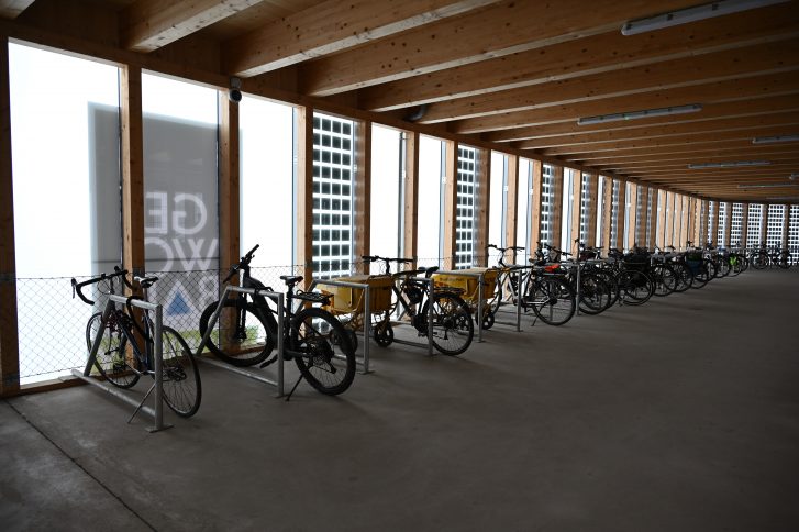 Innenraum des Fahrradparkhaus. Holzdecke, Betonbode, Anlehnbügel, an denen Fahrräder angeschlossen sind. insgesamt wirkt der Raum sehr ordentlich, hell und offen. 