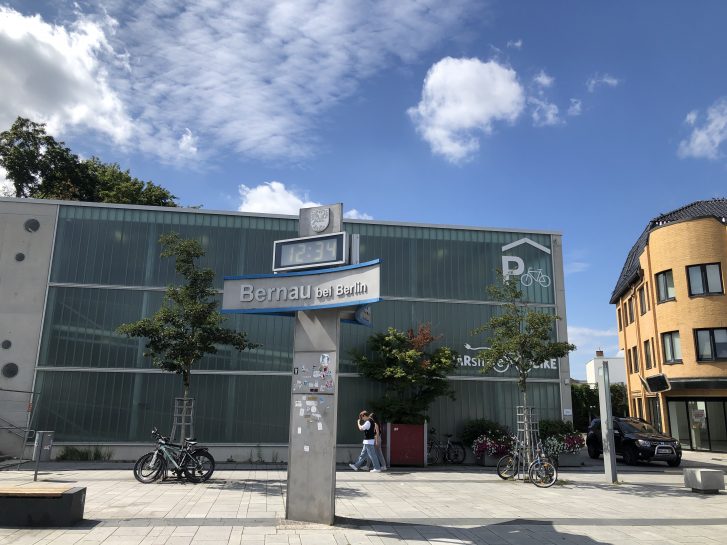 Öffentlicher Platz in Bernau bei Berlin. Im Vordergrund befindet sich eine Säule mit einer Uhr, die 12:34 Uhr anzeigt, und einem Schild mit der Aufschrift 