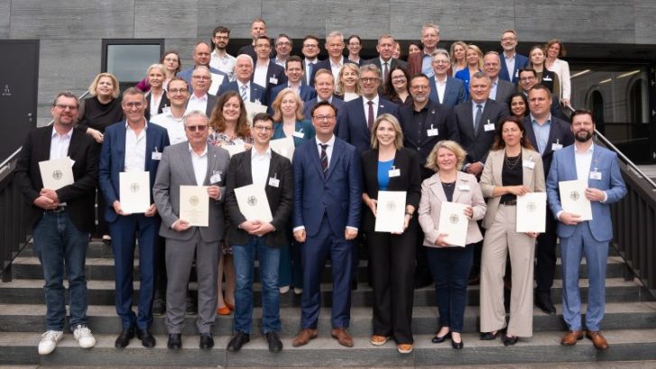 Menschen stehen auf einer Treppe für ein Gruppenbild aufgereiht und halten weiße Papierurkunden in den Händen