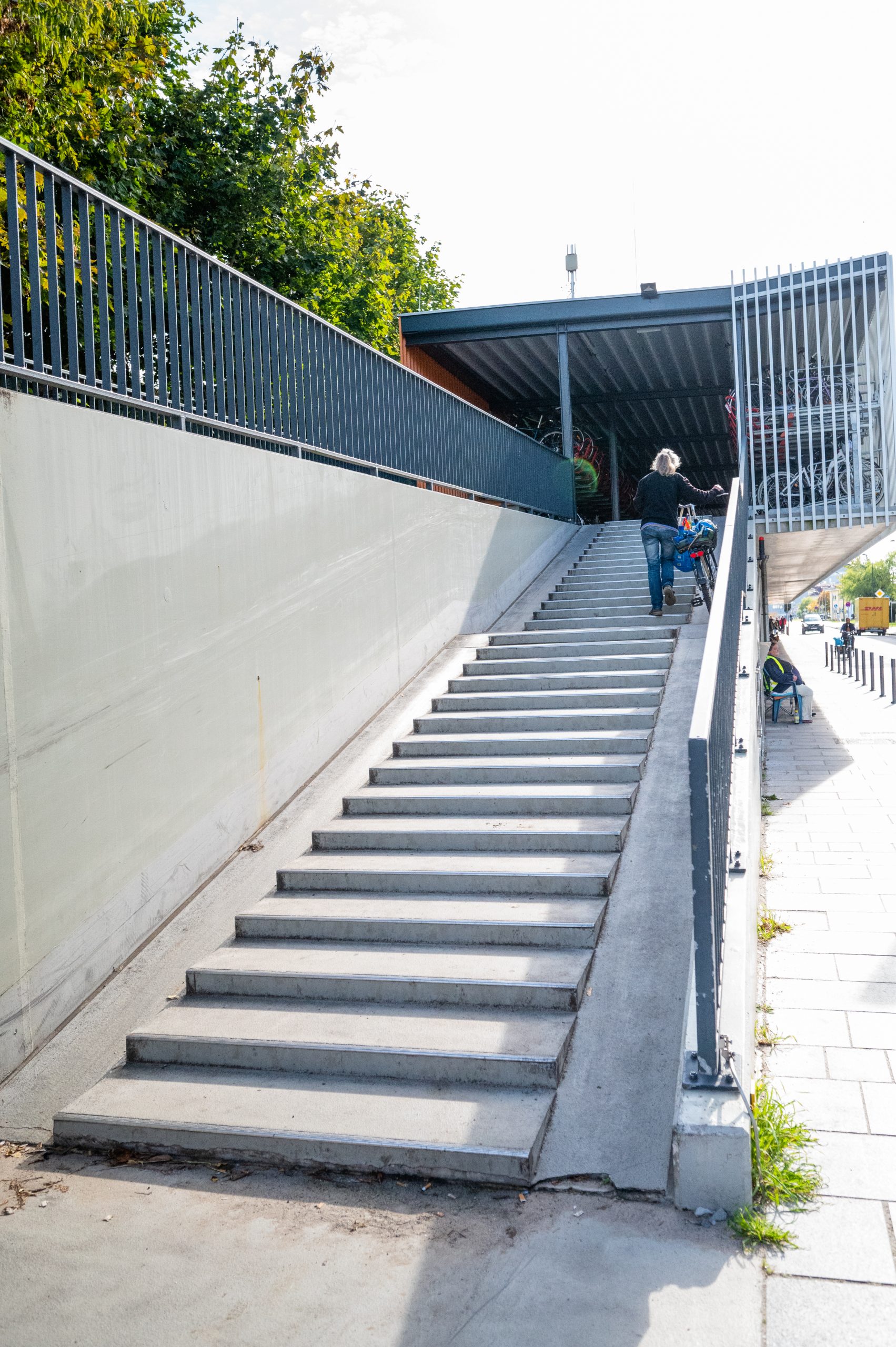 Eine Frau schiebt ihr Fahrrad an der Außenseite des Fahrradparkhauses auf betonierten Schieberinnen neben der Treppe vom EG ins OG hinauf 