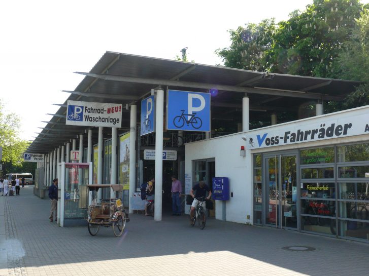 Gebäude mit mehreren Schildern, darunter "Fahrrad-Waschanlage", "Fahrradverleih", und "Voss-Fahrräder". Vor dem Gebäude parken Fahrräder und ein Mann fährt gerade mit einem Fahrrad weg.