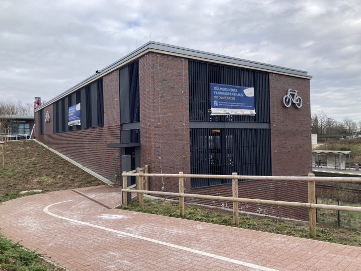 Blick auf die Zuwegung in Richtung Fahrradgarage, die aus roten Klinkersteinen besteht. EIn Fahrradweg endet direkt im Fahrradparkhaus. Der Fahrradweg ist durch einen Holzzaun begrenzt.