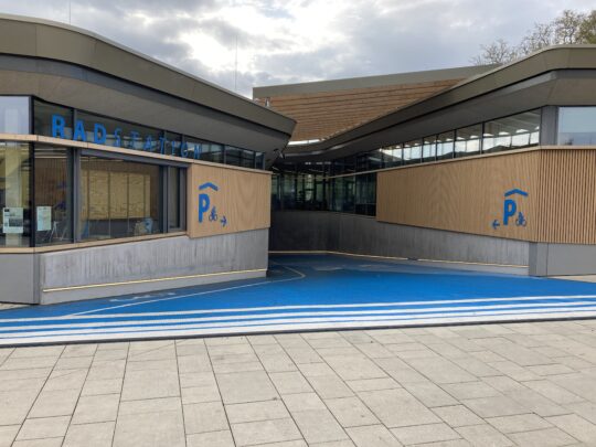 Eine blau-eingefärbte Rampe führt zwischen zwei Gebäudeteilen in das Untergeschoss der Radstation in Tübingen.