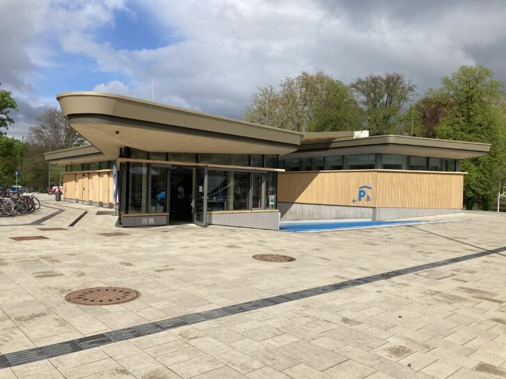 Außenansicht der Radstation Tübingen. Ein aus Holz gebautes Gebäude mit Glasfront. Eine blau-eingefärbte Rampe führt in das Untergeschoss. 