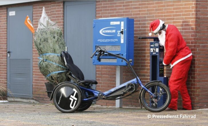 Weihnachtsmann am Schwalbe Schlauchautomat pumpt Lastenrad auf, in dem ein Weihnachtsbaum steht