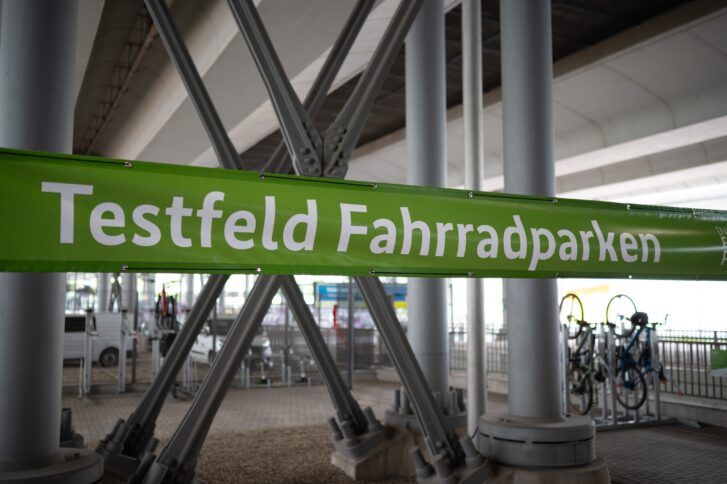 Grüner Banner mit weißer Schrift " Testfeld Fahrradparken", der unter einem Zugviadukt zwischen zwei Pfeilern hängt.