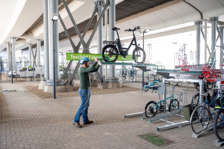 Eine Person schiebt ein Fahrrad auf die obere Schiene eines Doppelstockparkers, um es ordnungsgemäß zu parken. Im Hintergrund ist ein hellgrünes Banner, auf dem 'Testfeld Fahrradparken' steht, sichtbar. Der Boden besteht aus grau gepflasterten Steinen, und oberhalb des Testfeldes sind die Bahngleise.