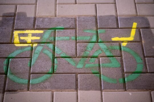 Bodenmarkierung mit Kreidespray. Es zeigt ein Fahrrad in grüner Farbe. Am Fahrrad sind in gelber Farbe ein Korb und ein Kindersitz sichtbar.