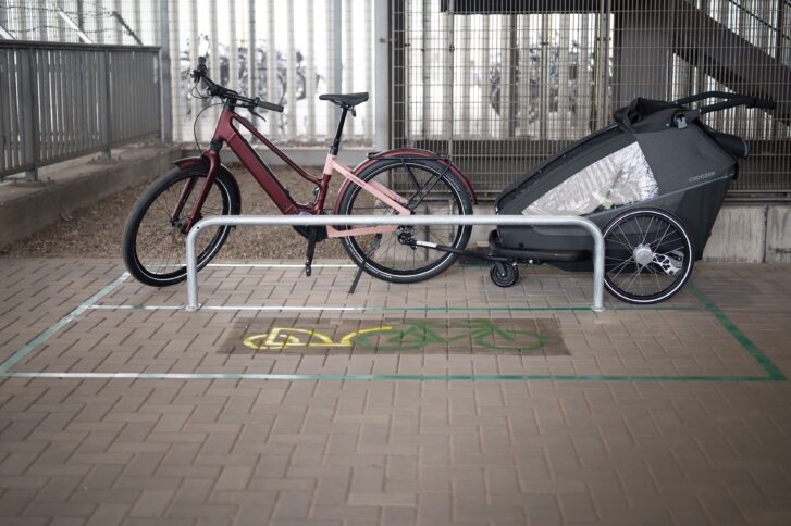 Ein niedriger und länglicher Fahrradbügel, an dem ein Fahrrad mit Anhänger geparkt ist. Auf dem grauen Pflaster vor dem Bügel befindet sich ein Piktogramm mit einem grünen Fahrrad und einem gelben Anhänger.