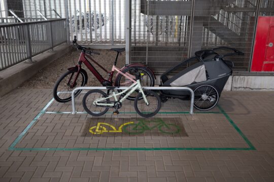 Ein Fahrrad mit Kinderanhänger sowie ein Kinderfahrrad lehnen an einem Fahrradbügel, der auf einer Fläche steht, auf der ein Piktogramm eines Rades mit Anhänger sichtbar ist.