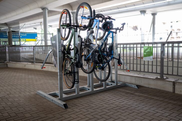 Ein Senkrechtparker, bei dem Fahrräder am Arm mit dem Vorderrad eingehängt werden können. Er ist beidseitig bedienbar und bietet Platz für 10 Fahrräder. Derzeit sind drei Fahrräder geparkt. Die Fahrradabstellanlage befindet sich unter einem Bahnviadukt. Der Boden ist gepflastert und hinter der Anlage befindet sich ein Geländer.