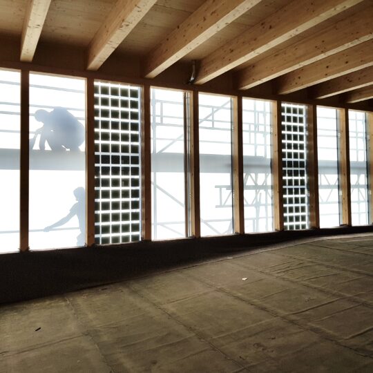 Innenansicht eines im Bau befindlichen Fahrradparkhauses mit Holzdeckenbalken. Durch große vertikale Fensteröffnungen sind Gerüste und die Silhouetten von Bauarbeitenden zu sehen