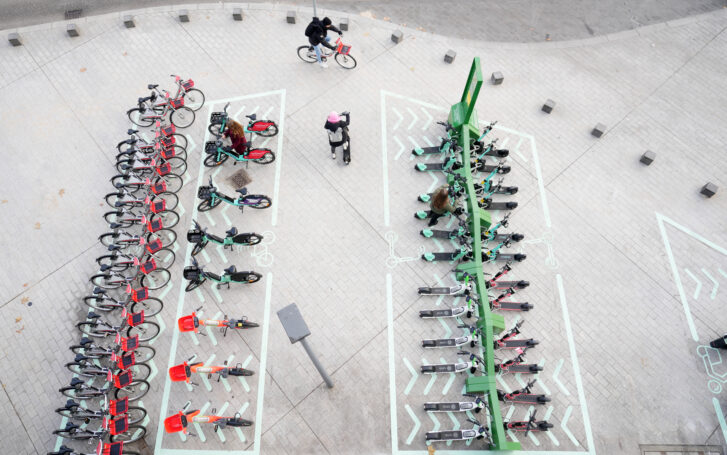 Luftaufnahme eines städtischen Mobilitätshubs mit geordnet abgestellten Leihfahrrädern und E-Scootern in markierten Parkflächen; mehrere Personen bewegen sich mit Fahrrädern und Scootern über den gepflasterten Platz.