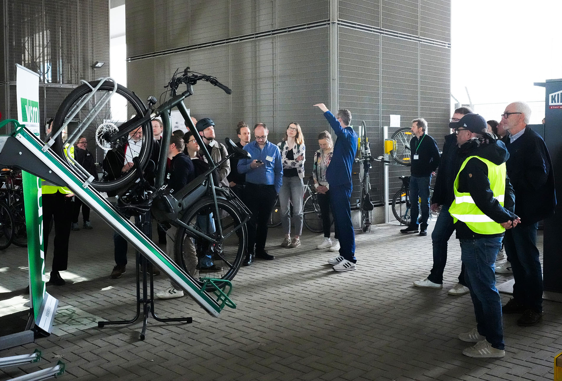 Eine Gruppe von Konferenzteilnehmenden betrachtet ein auf einer Schräge montiertes Lastenrad auf einem Fahrradparkständer. Ein Mann in blauer Jacke erläutert die Anlage.