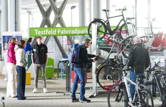 Innenaufnahme einer Fachmesse zum Thema Fahrradparken. Ein grünes Banner mit der Aufschrift ‚Testfeld Fahrradpark[en]' ist im Hintergrund zu sehen. Rechts im Bild befindet sich ein mehrstöckiges Fahrradparkgestell (Doppelstockparker) mit mehreren eingeparkten Fahrrädern, darunter E-Bikes und Lastenräder. Ein Mann mit blauem Rucksack bedient das System, eine weitere Person steht daneben. Links im Bild unterhalten sich drei Personen in der Nähe eines Messestands